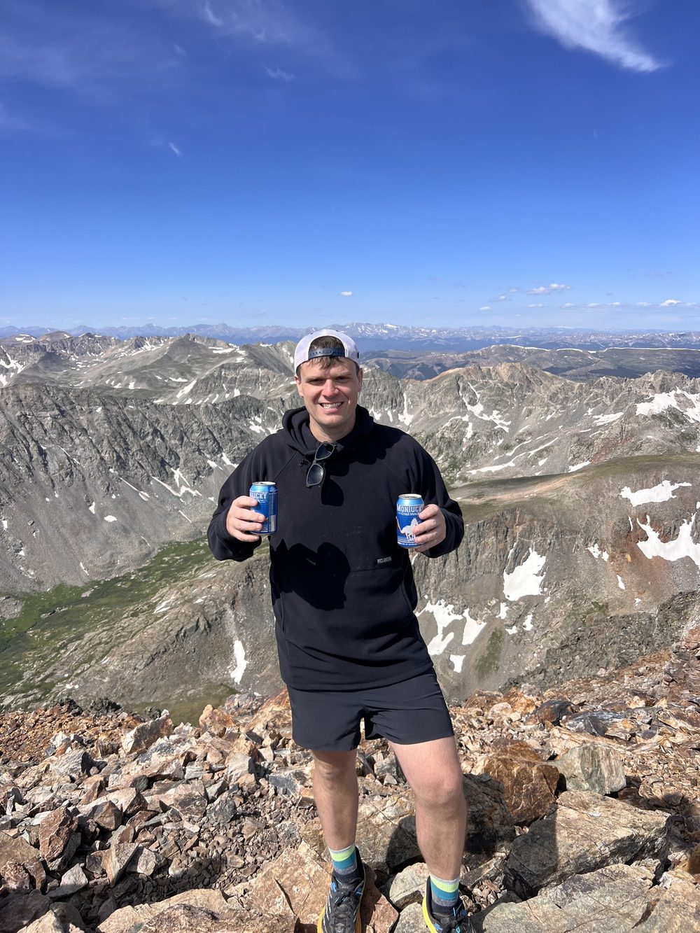 Josh Shelley smiling on a Colorado mountain summit, holding two beers